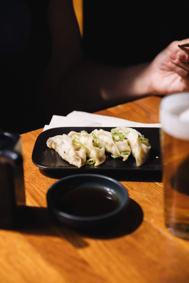 La imagen presenta una escena íntima y cálida en un restaurante. En el primer plano, hay un plato negro rectangular sobre una mesa de madera que contiene seis gyozas cuidadosamente dispuestas y adornadas con finas rodajas de cebolla verde. Las gyozas tienen una apariencia dorada y tentadora, sugiriendo que están recién cocidas y posiblemente crujientes al tacto. A la derecha del plato, un pequeño cuenco de salsa de soja invita a sumergir las dumplings antes de saborearlas. También se ve un vaso de cerveza con espuma, lo que añade un toque de relajación a la atmósfera. La luz suave y la estrecha profundidad de campo centran la atención en la comida, mientras que en el fondo, la presencia difuminada de una persona con una blusa amarilla y un gesto tranquilo de las manos añade un elemento humano a la escena, sugiriendo la posibilidad de una conversación amena o una pausa reflexiva durante la comida. en 4 makis